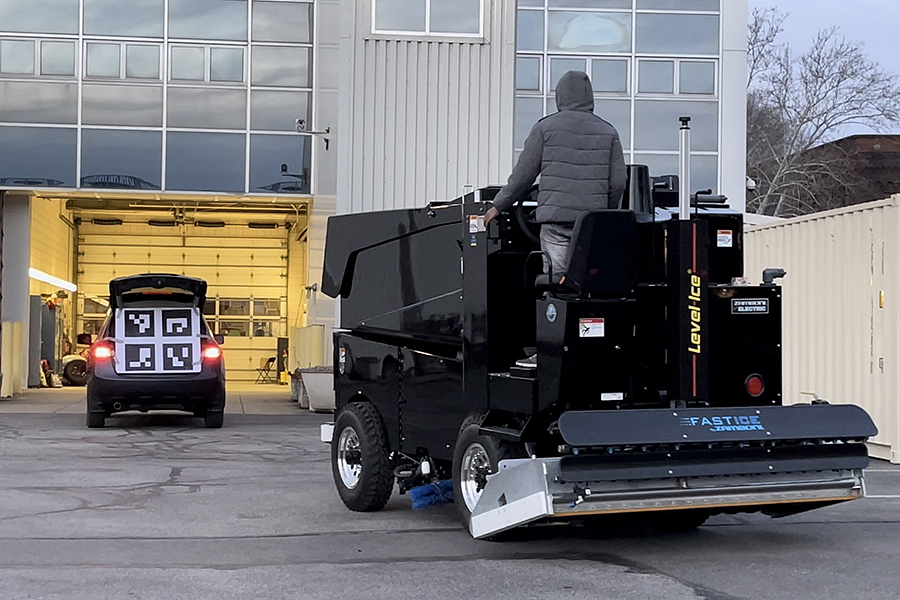 A Zamboni machine follows an SUV with a large code resembling a QR code on its liftgate outside a large industrial-looking garage.