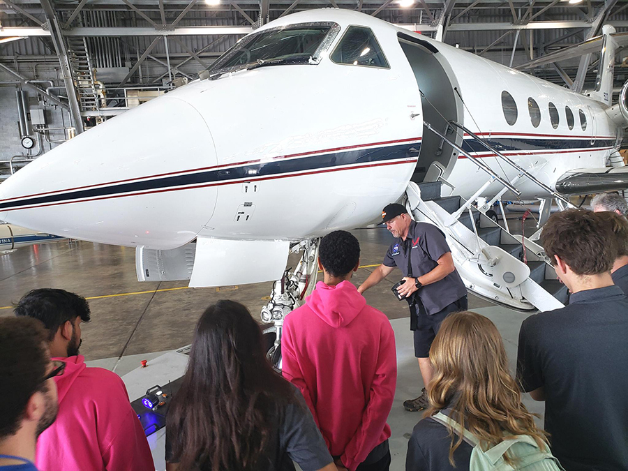 Students with their backs to the camera look at a small airplane in a hanger. A man in dark shorts, a button-down short-sleeved shirt and a hat points to an area near the plane's wheel.