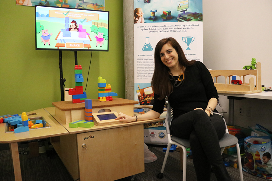 A woman interacts with stacked blocks on a table. Behind the blocks, a computer screen shows a cartoon gorilla and two pigs offering tips and advice for building strong structures.