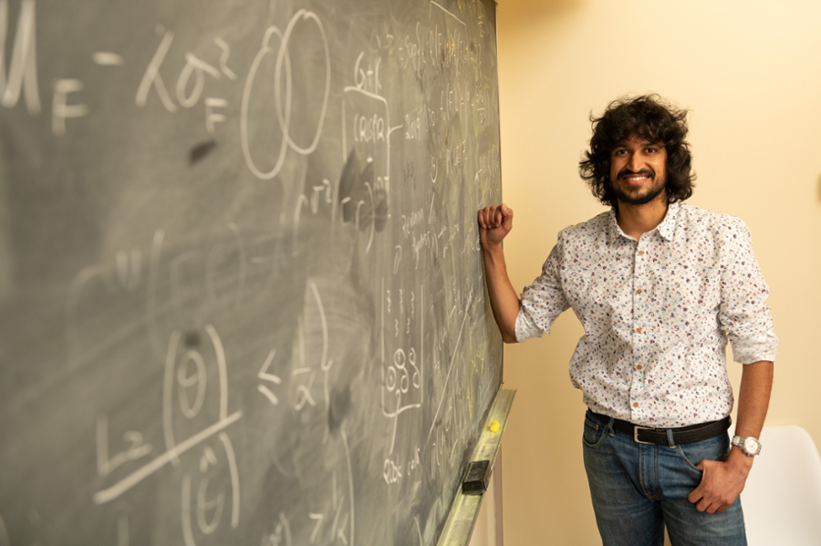 Aaditya Ramdas stands to the right of a black chalkboard covered in scientific formulas.