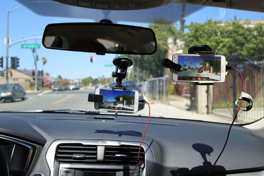 Two cell phones hang from the inside of a car windshield near the review mirror with the dashboard at the bottom of the shot.