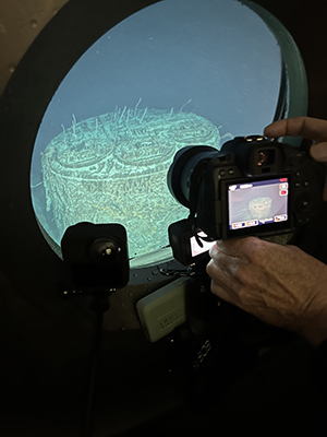 A photo of a boiler on the Titanic. The boiler is seen through the porthole of a submersible during a dive. The boiler is covered in a green algae. 