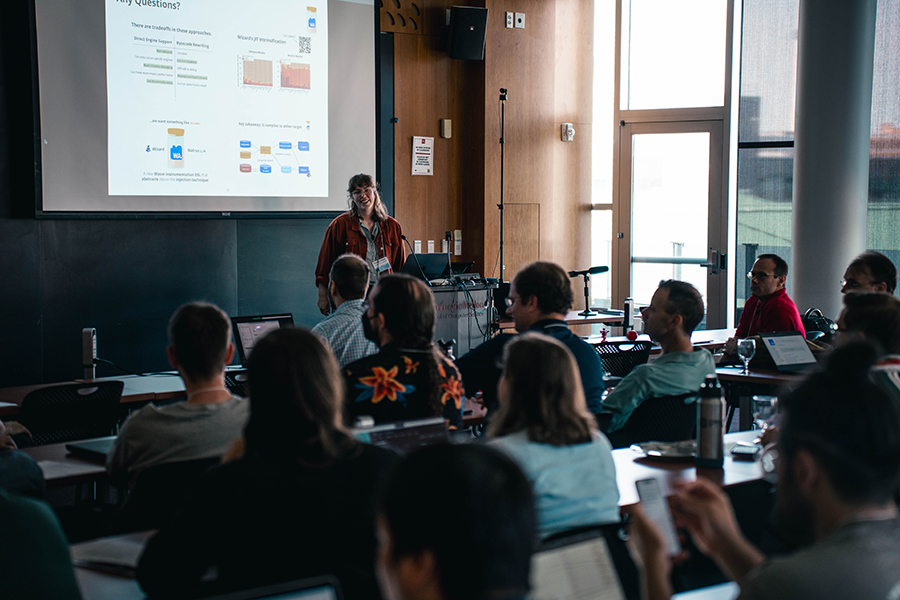A woman with brown hair stands behind a podium in a classroom while a screen behind her displays talk slides.